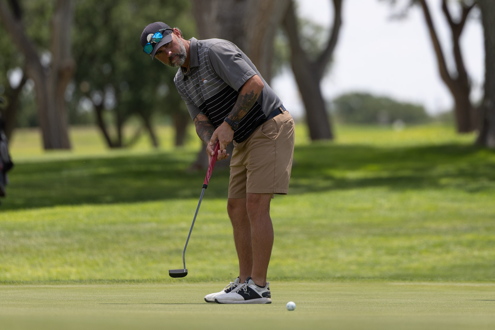 Golfer bending to line up a putt on a lush green course, wearing a cap and striped polo.