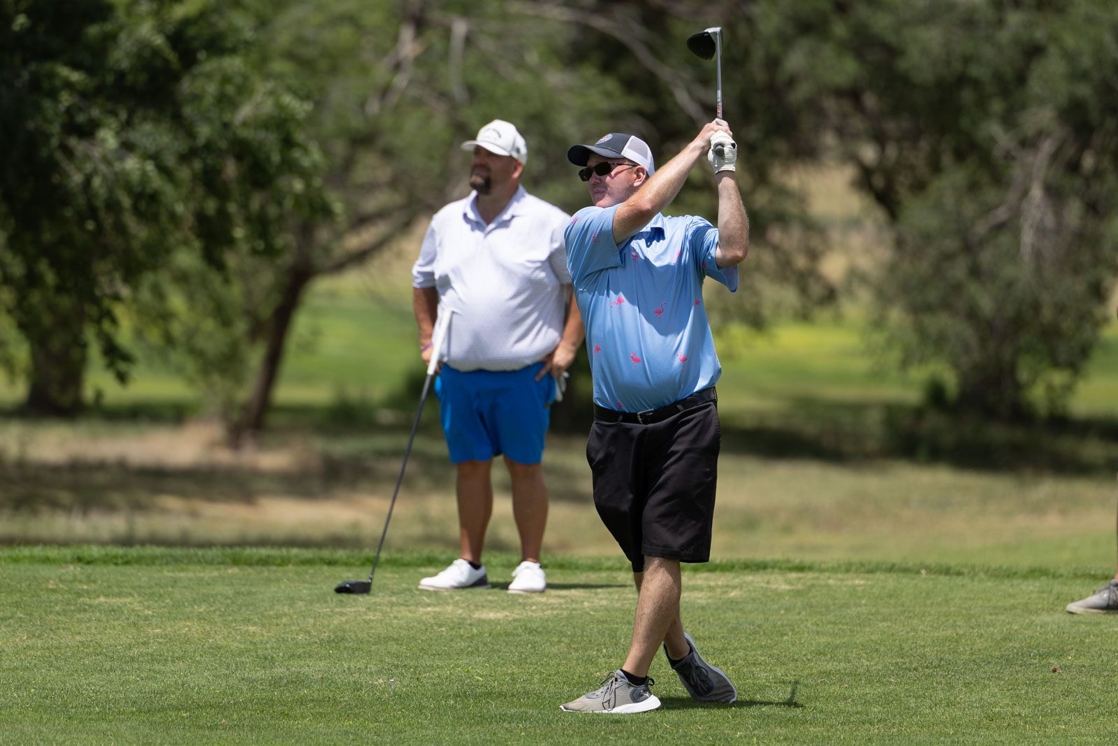 Golfer in a blue shirt mid-swing on a sunny golf course, watching companion in white nearby.
