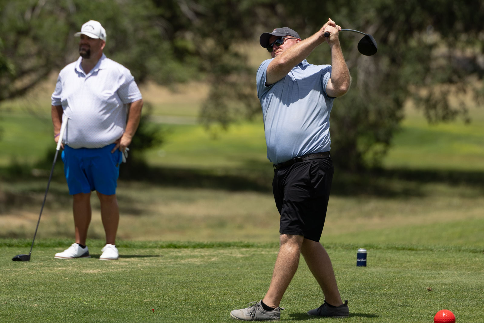 Two men on a golf course: one swings a club while the other watches nearby.
