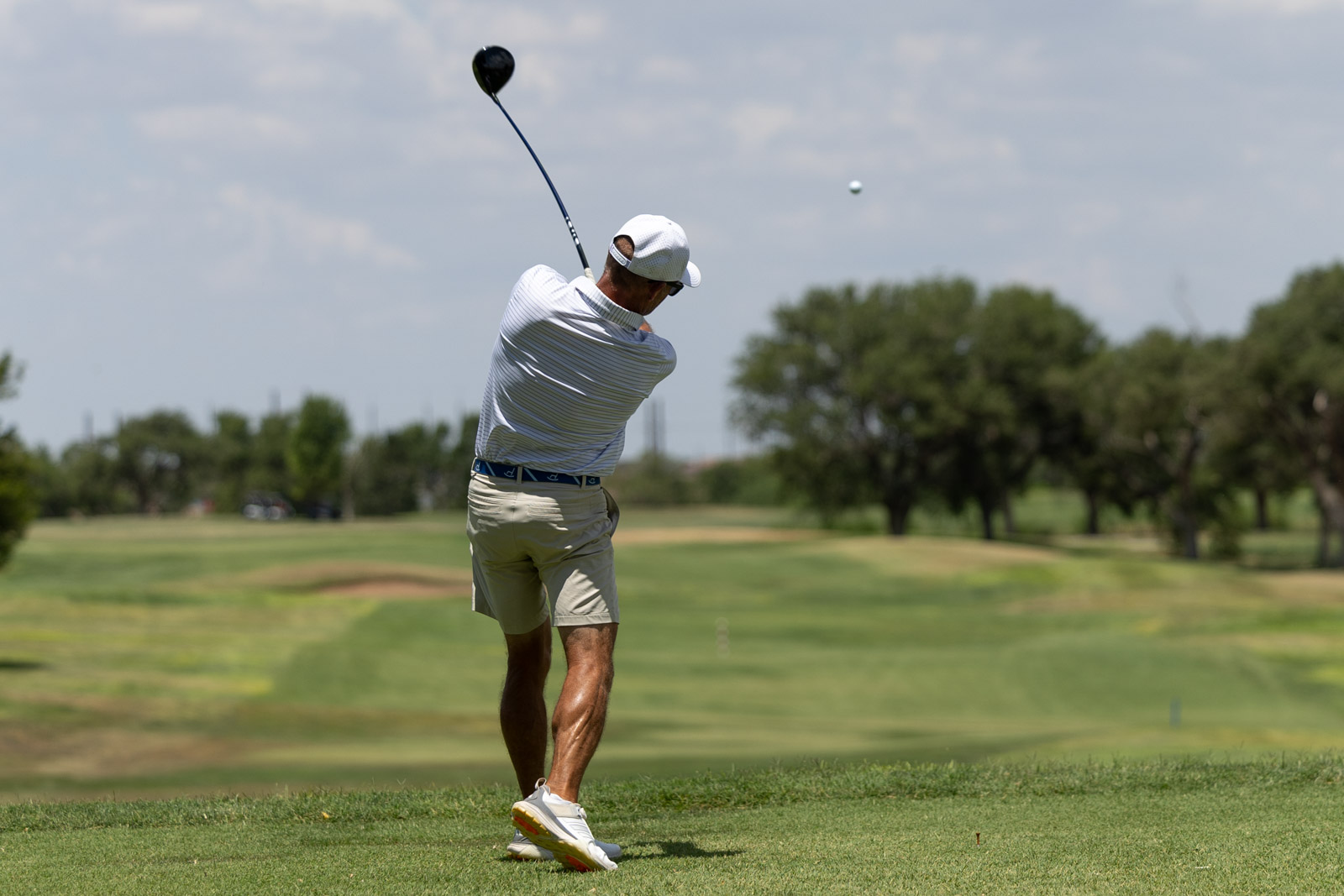 Golfer in a white cap and striped shirt mid-swing on a sunny golf course; golf ball in flight to the right.