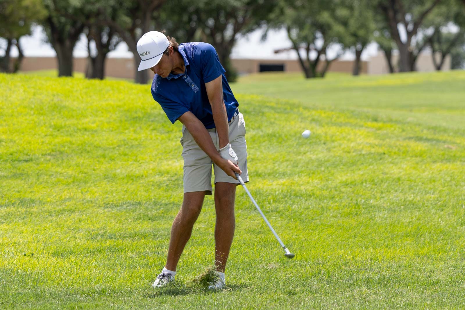 Golfer in blue shirt and white cap swinging a club on a sunny golf course.