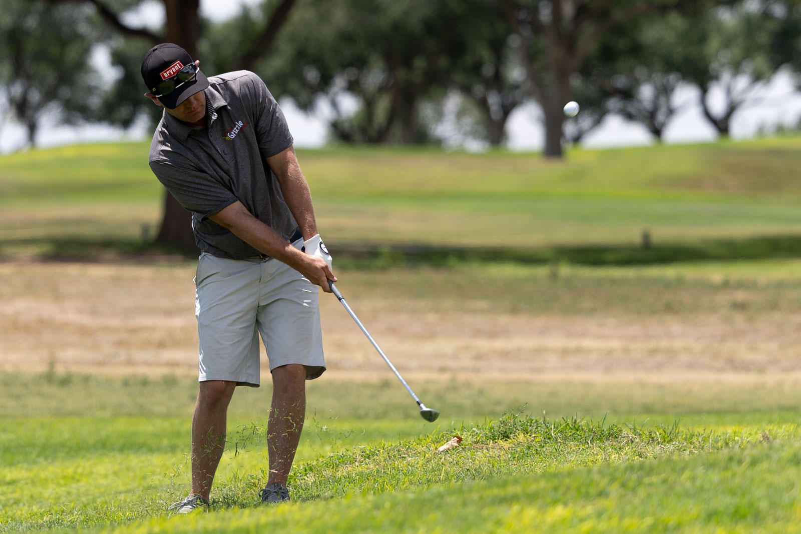 Man in a dark cap and grey polo tees off on a sunny golf course, mid-swing with a driver.