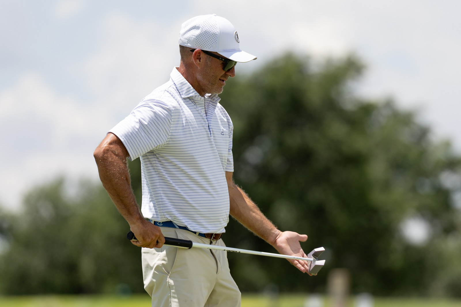 Older man in a white striped polo and cap holding a golf club on a sunny golf course.