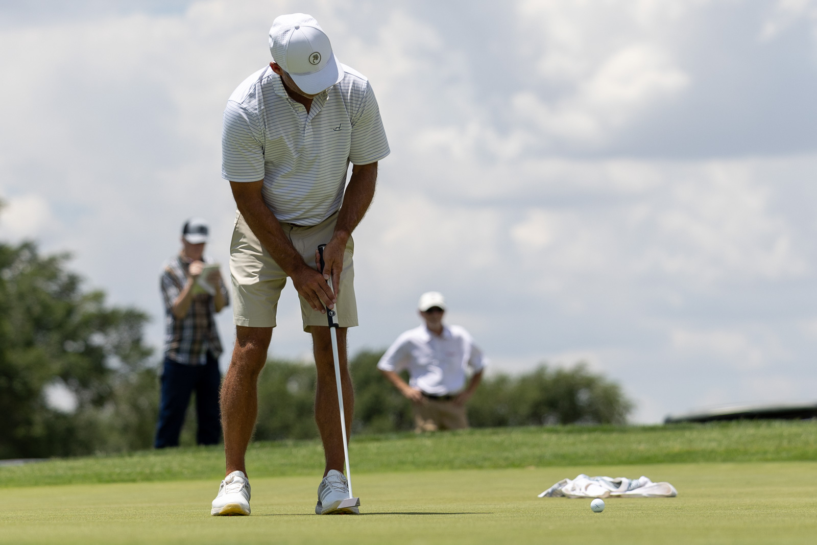 Golfer lining up a putt on the green with two spectators watching in the background on a sunny course.