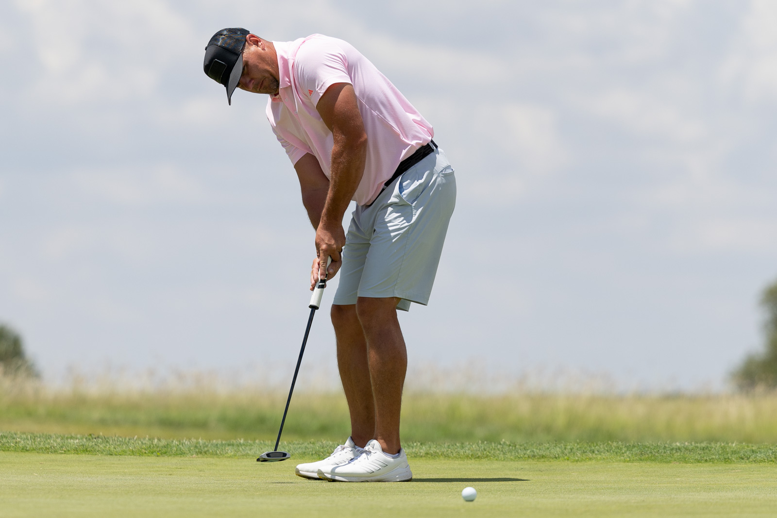 Golfer in a pink polo shirt and light gray shorts lines up a putt on the green under a sunny sky.