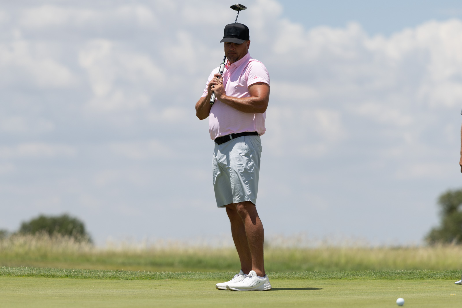 Golfer in a pink polo and black cap stands on a putting green, holding a club near his mouth with a golf ball visible on the grass nearby.