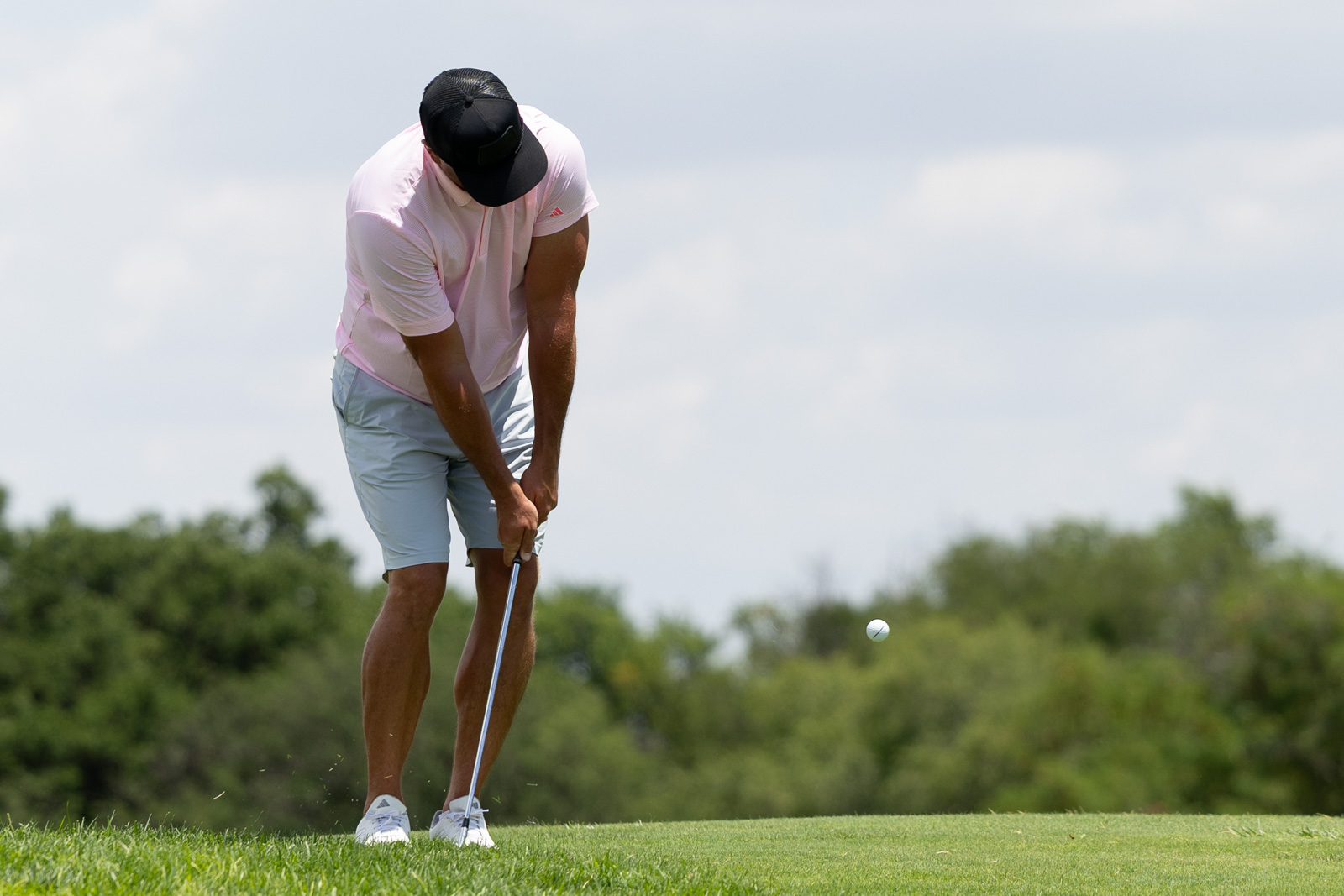 Golfer in a pink shirt and black cap lines up a putt on a sunny green course.