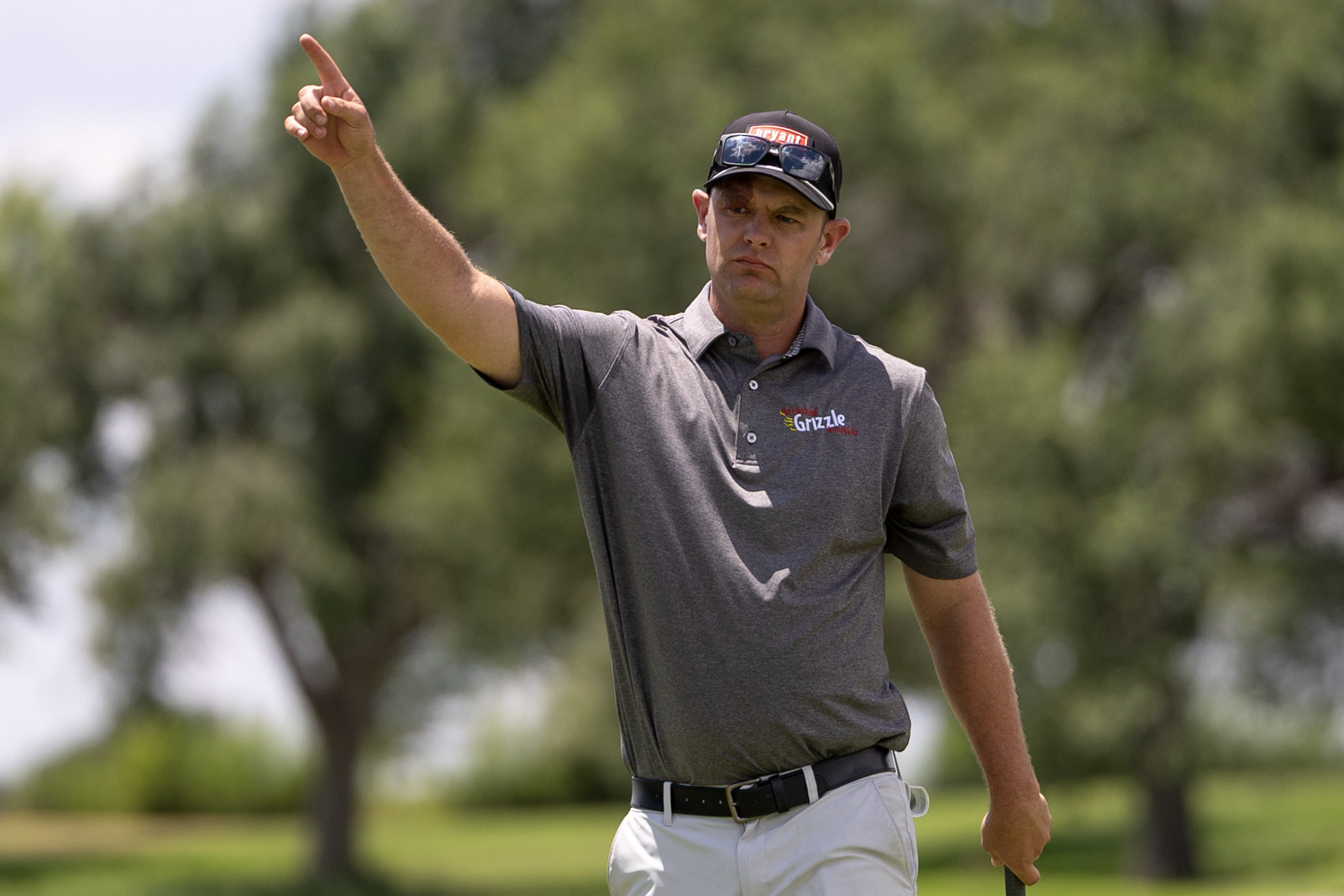 Golfer in a gray polo shirt and white pants points upward with his right arm on a sunny golf course.