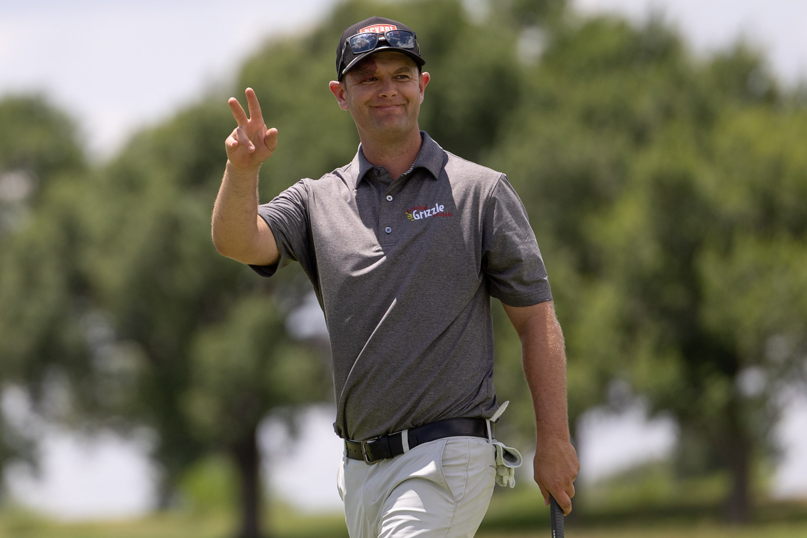 TheBud-38 | Press Pass Sports Man in gray polo and white shorts on a golf course, flashing a peace sign with his left hand