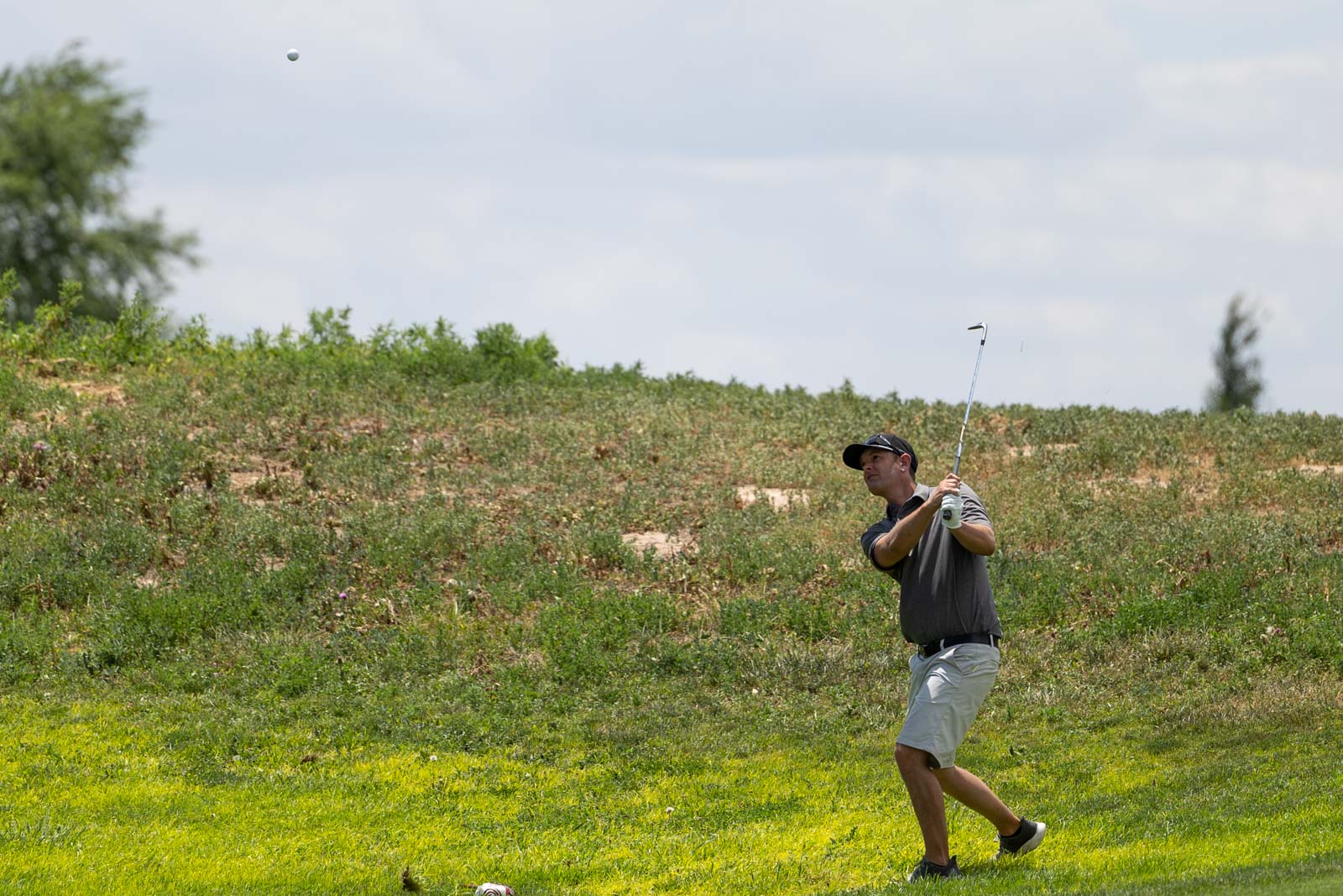 Golfer in a gray polo and cap swings a club on a grassy hill as the ball sails into the air.