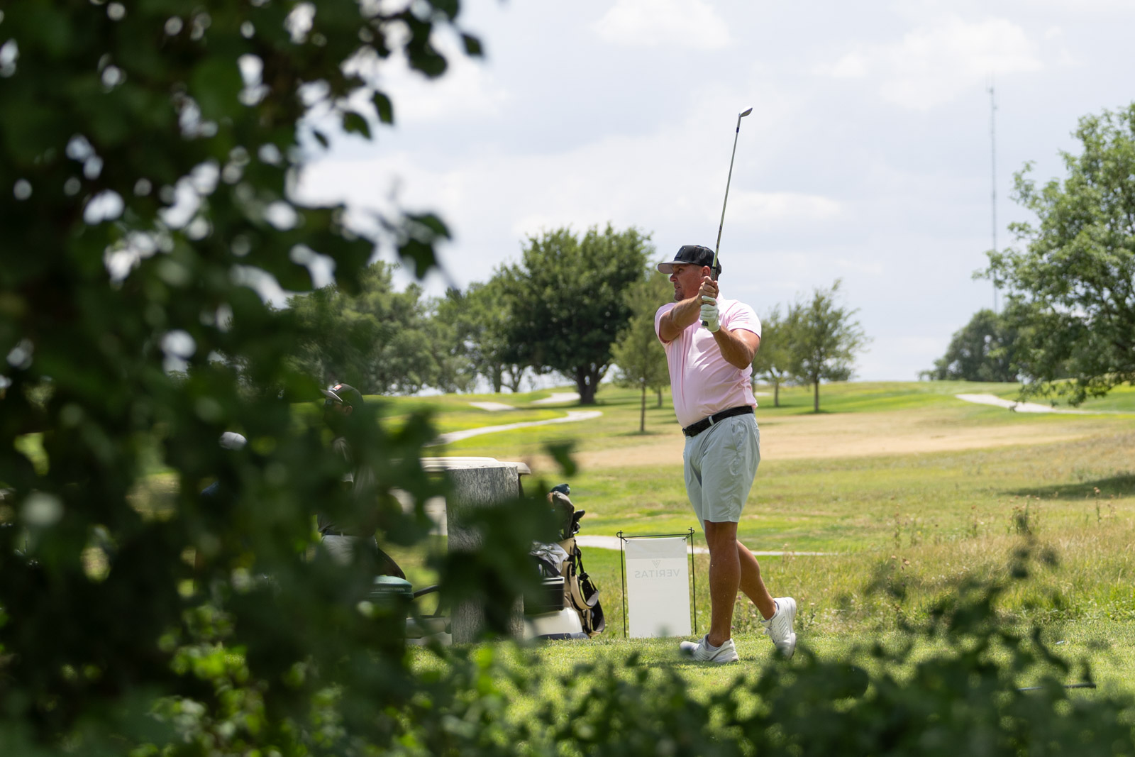 A man in a pink polo shirt and gray shorts mid-swing on a sunny golf course with trees in the distance.
