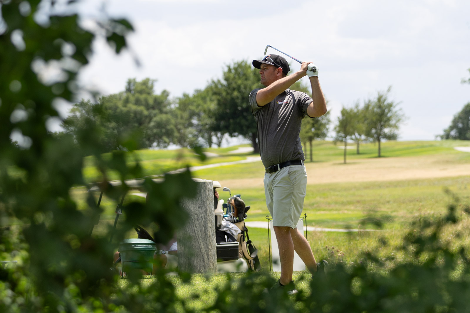 A man in a gray polo and cap swings a golf club on a sunny course, with trees in the background and a bag of clubs nearby.