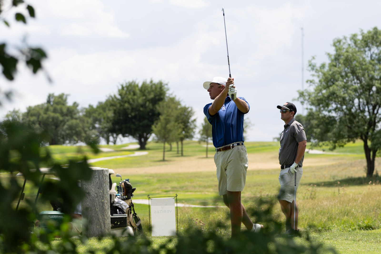 Two golfers on a sunny course: one in a blue shirt mid-swing, the other watching nearby.