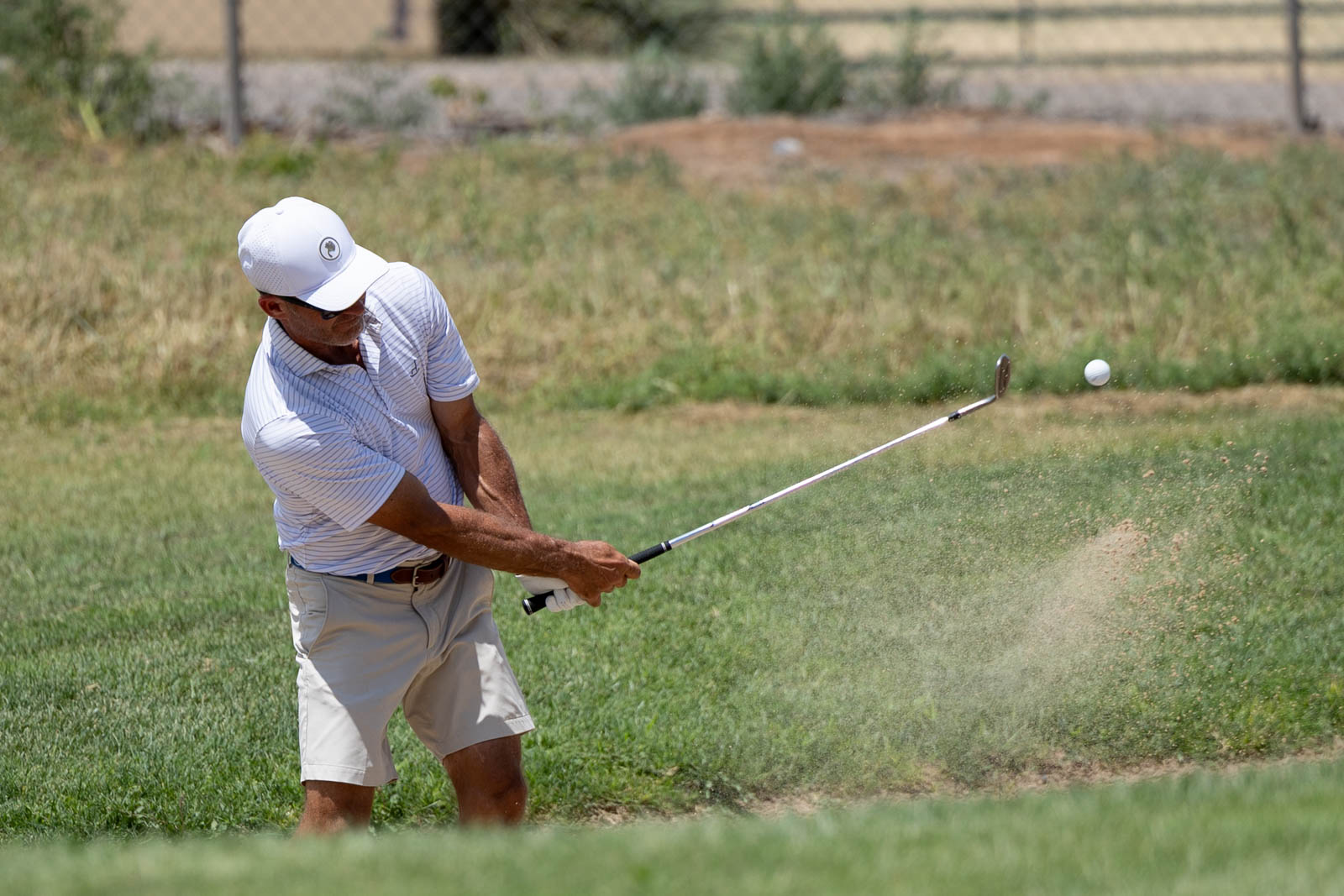 Golfer in a white cap and striped polo swinging at a golf ball on a sunny course.