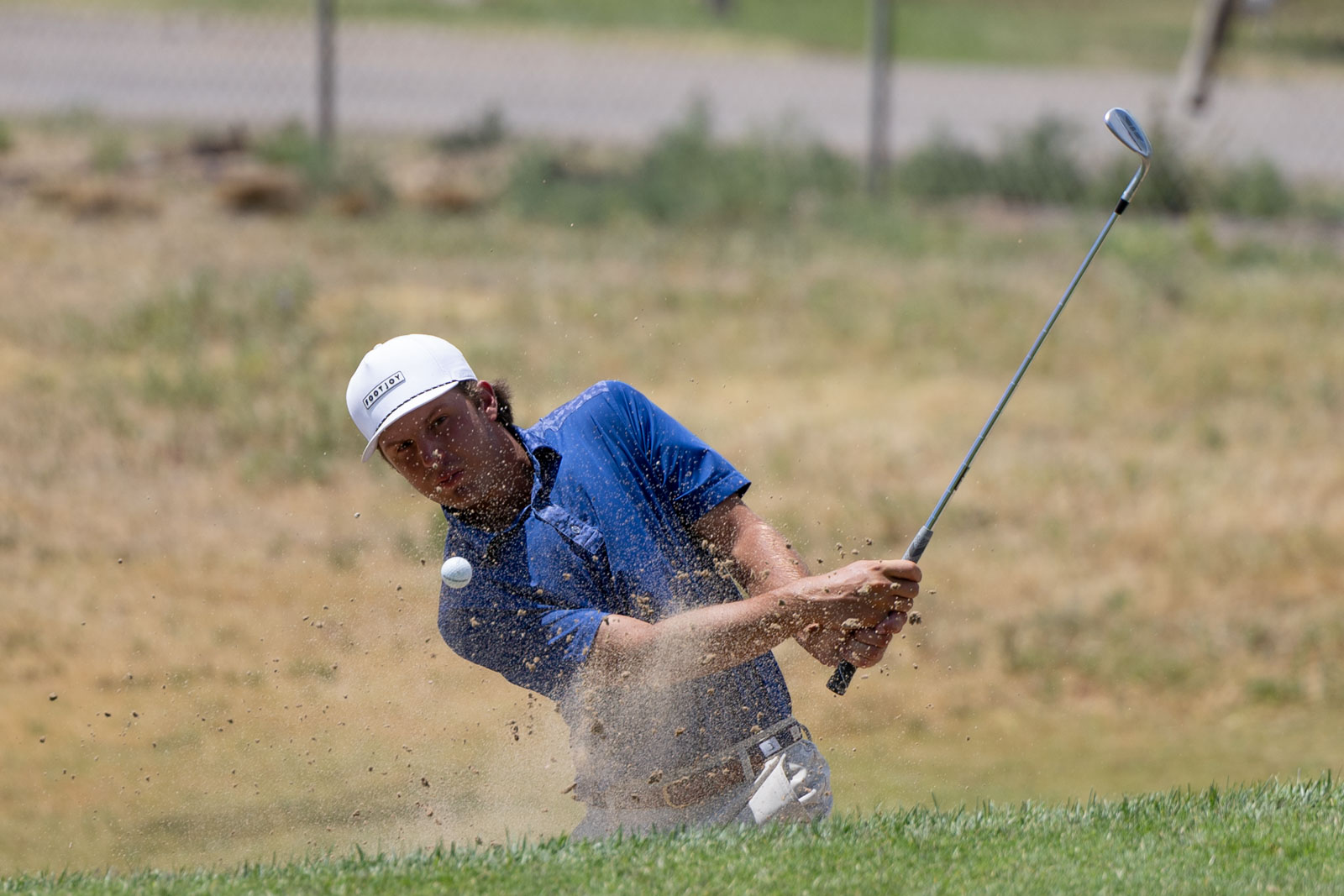 TheBud-48 | Press Pass Sports Golfer in a blue polo swinging from a sand bunker as sand flies up around him, with a white cap and golf club raised.