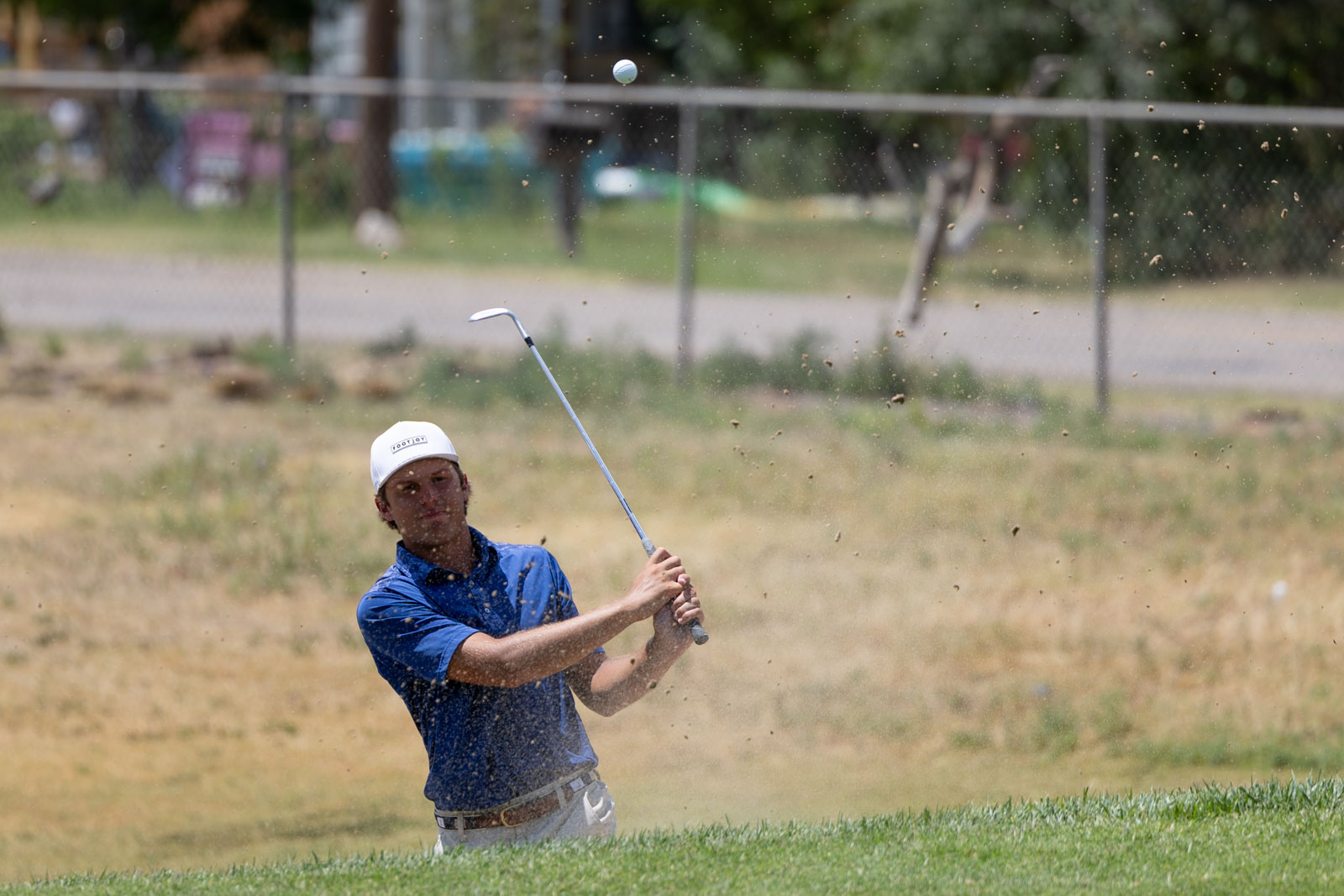 Golfer in a blue shirt and white cap hits a sand shot from a bunker as the ball rises and sand sprays around him.