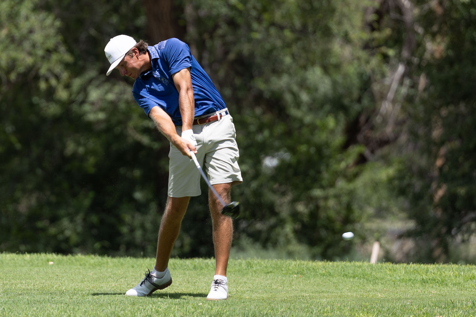 Golfer in blue shirt and white cap mid-swing on a green golf course.