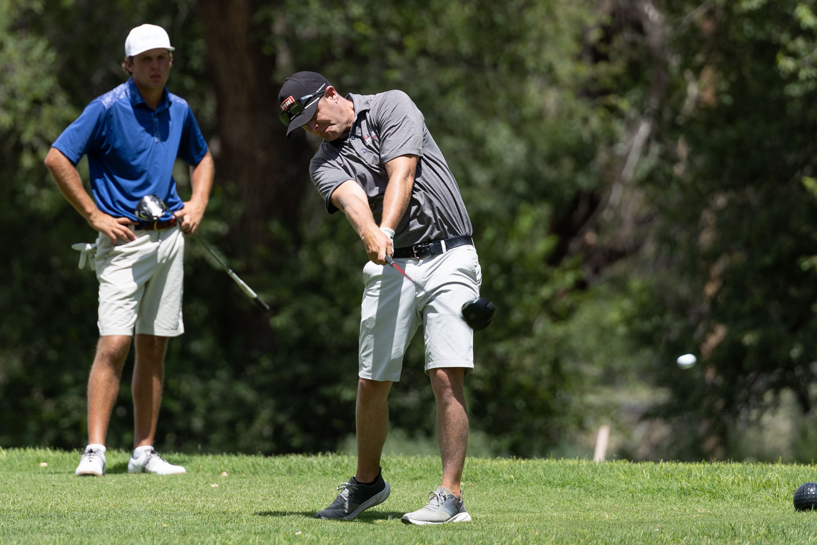 Two golfers on a sunny course: one man in a gray polo swinging a club as the other watches in a blue shirt and white cap on the grass.
