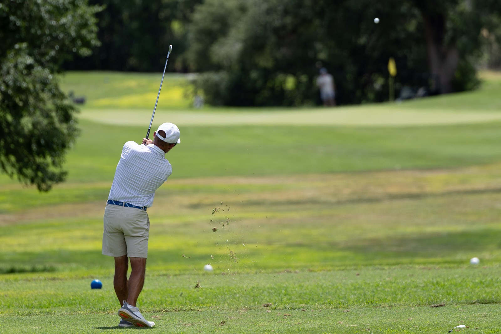 Golfer in a white cap and polo mid-swing on a sunny fairway, grass spraying from the impact of the ball.",