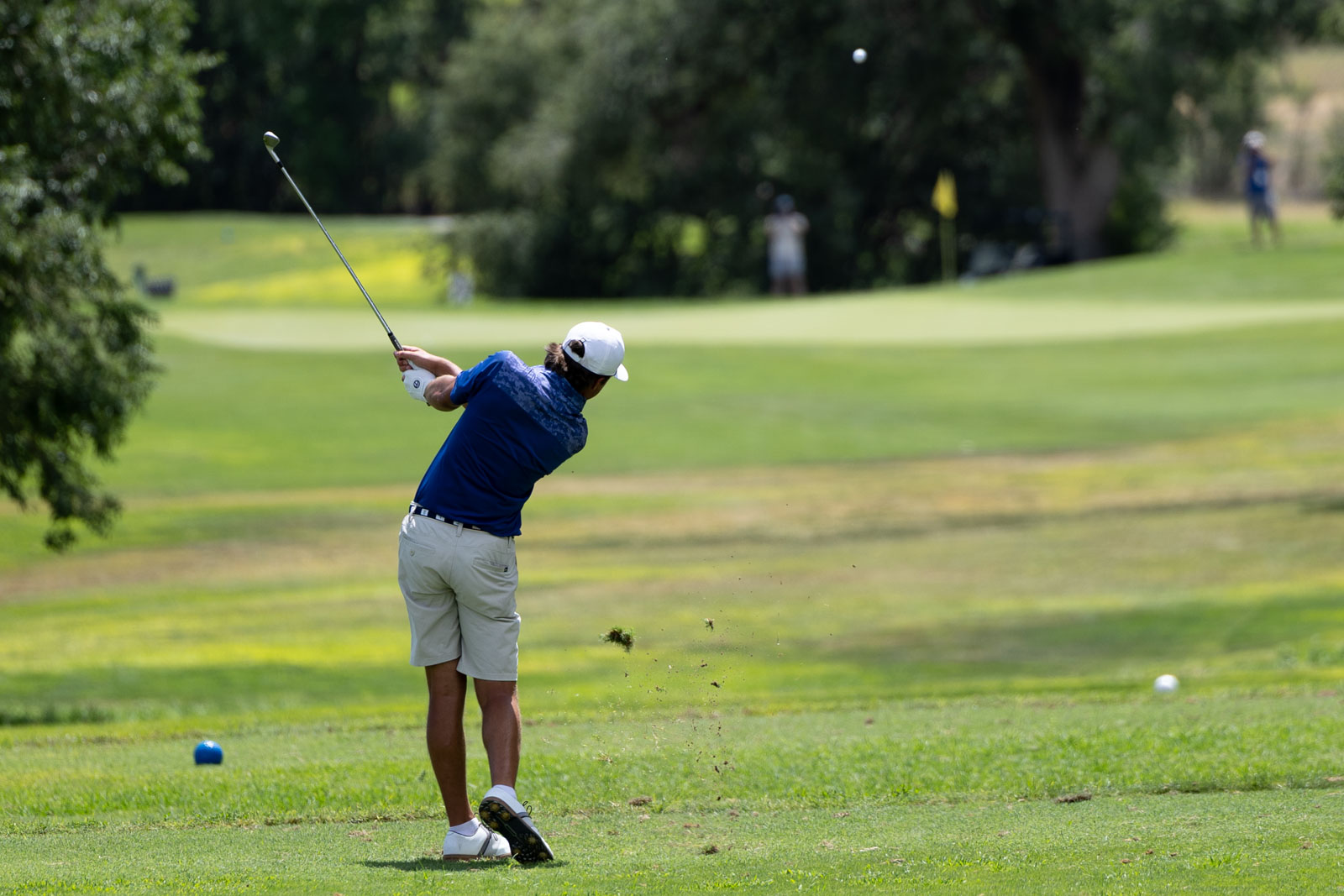Golfer in a blue shirt and white cap swinging a golf club on a sunny green course