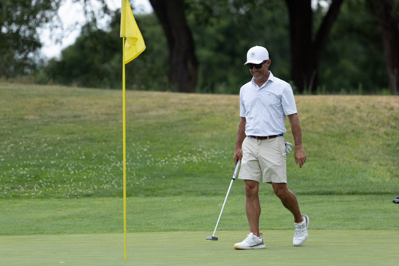 Golfer in a white striped polo, white cap and beige shorts walking on the green near a yellow flagstick by the hole, with trees in the background.