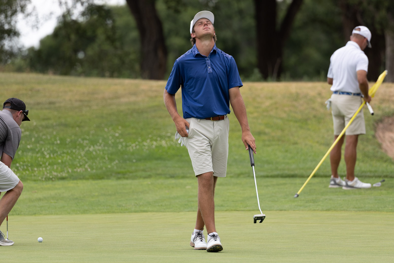 Golfer in a blue polo and white cap walks on the green, holding a putter as another player with a yellow flag stands in the background, preparing for a putt.