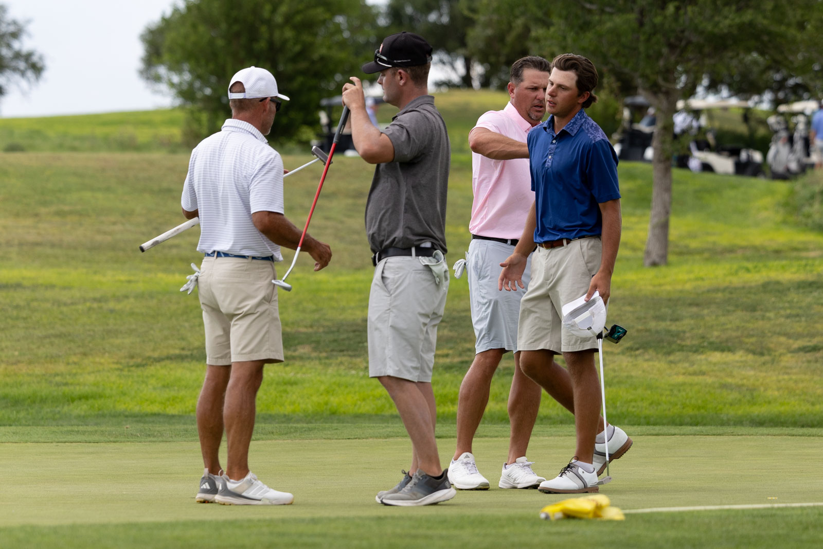 Group of four men on a golf course putting green, talking and holding clubs, with hats and golf gloves visible.