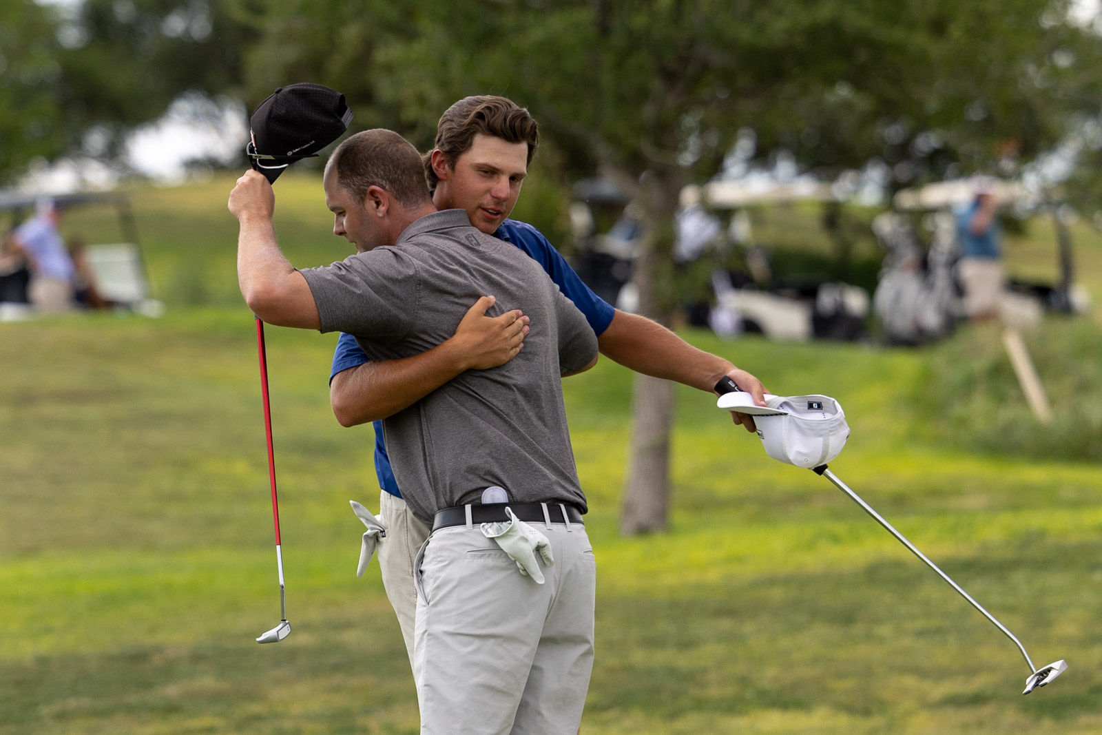 Two golfers embrace on a green at the golf course, one lifting a black cap in celebration while a club rests nearby on the ground.