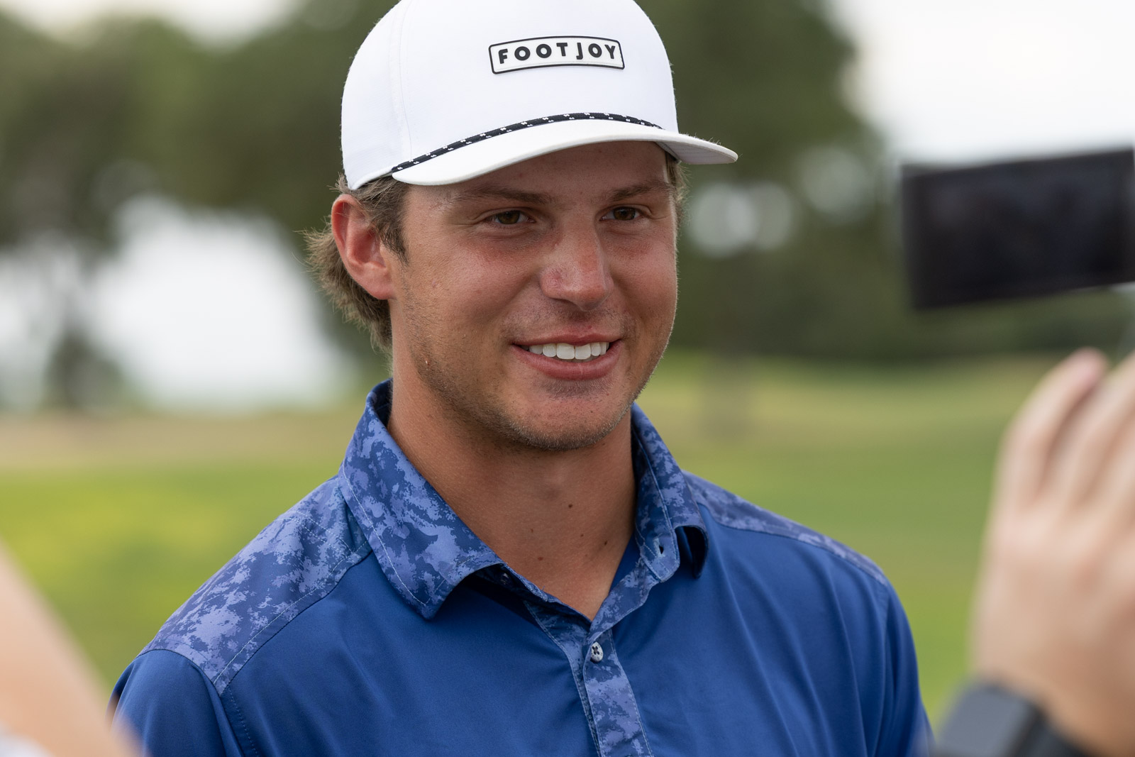 Smiling man in a white FootJoy cap and blue shirt being filmed outdoors (selfie context).
