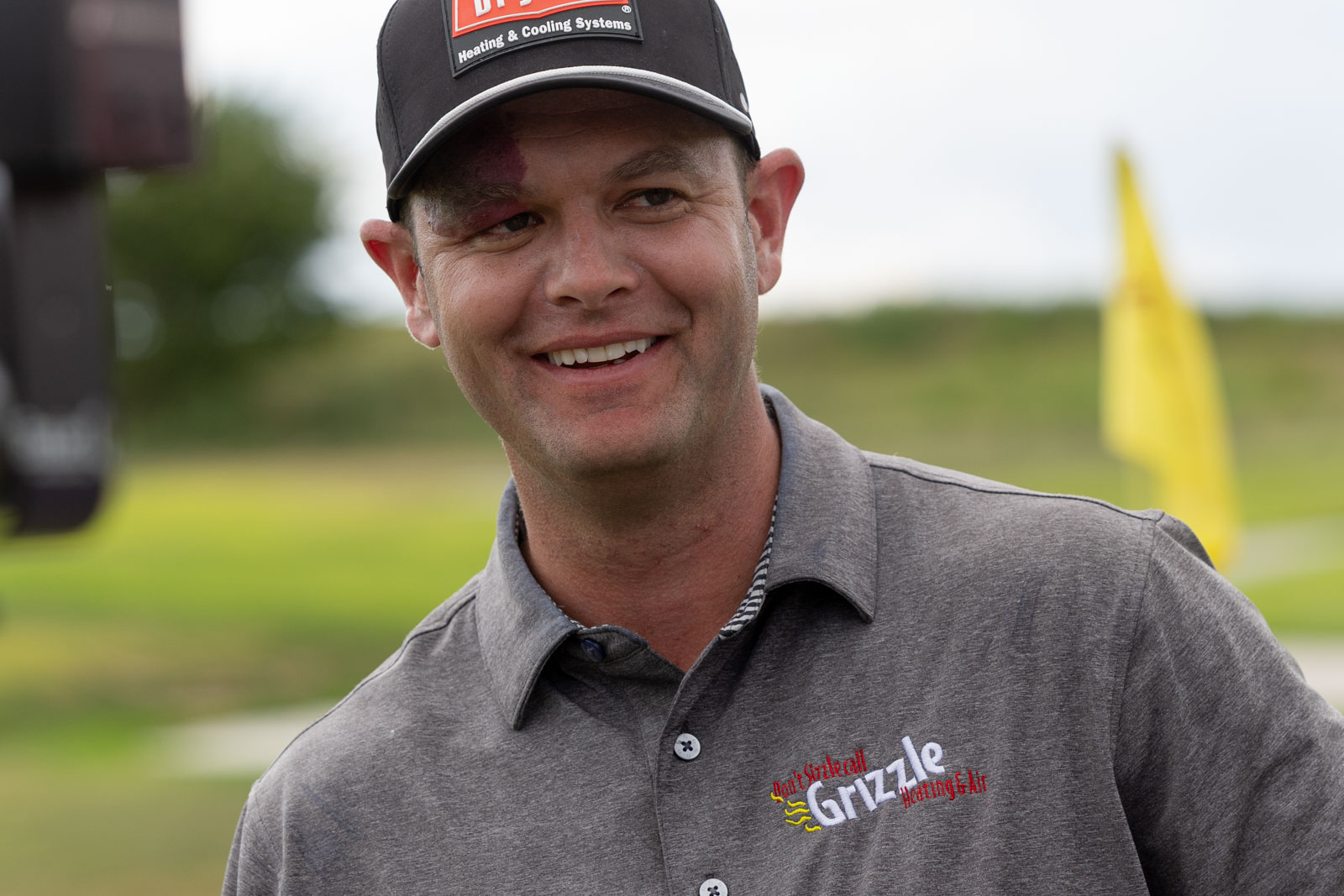 Smiling man wearing a black cap and gray polo shirt with a 'Grizzle Heating & Air' logo, outdoors in a grassy area.