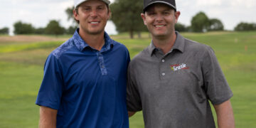 Two men stand side by side smiling on a golf course, wearing polo shirts and baseball caps with sponsor logos, greenField background.