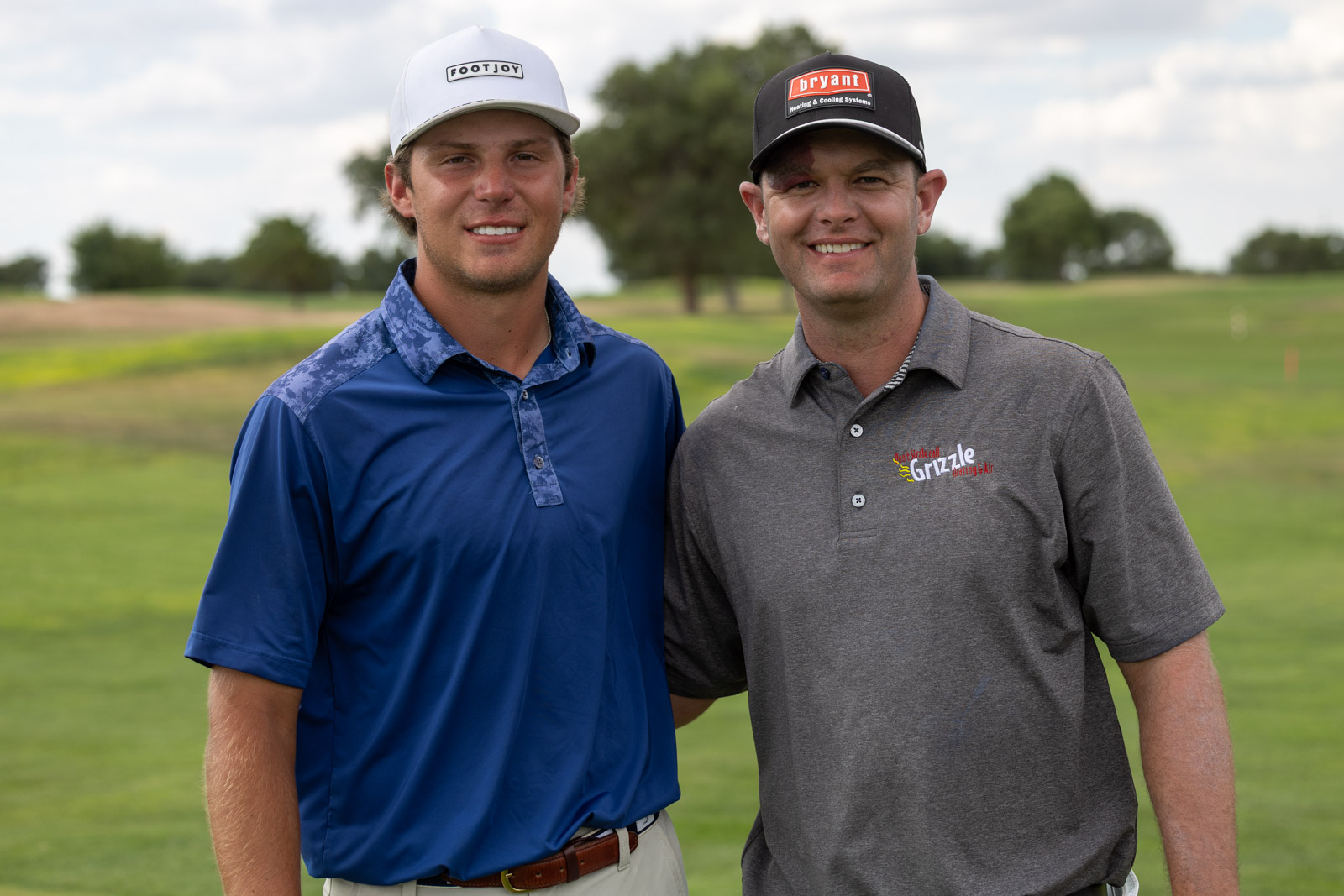 Two men stand side by side smiling on a golf course, wearing polo shirts and baseball caps with sponsor logos, greenField background.