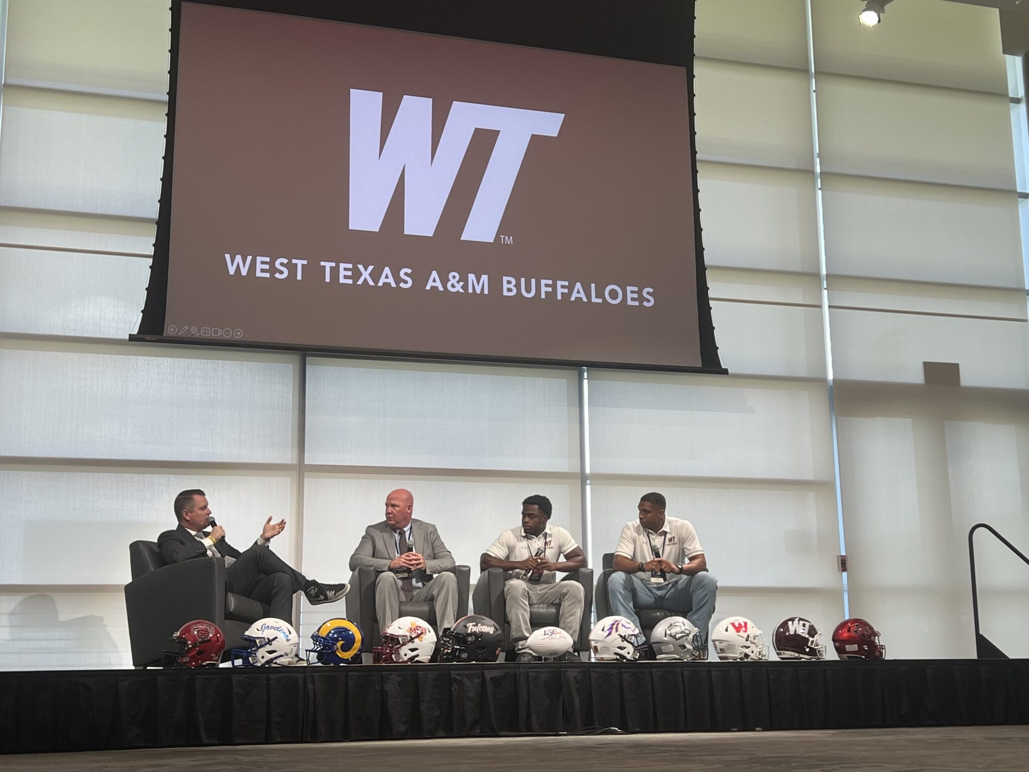 IMG_4305 | Press Pass Sports Panel discussion on a stage with four men in suits; football helmets lined up at the edge, WTAM Buffaloes logo on screen behind