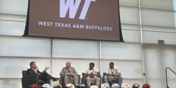 Panel discussion on a stage with four men in suits; football helmets lined up at the edge, WTAM Buffaloes logo on screen behind