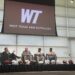 Panel discussion on a stage with four men in suits; football helmets lined up at the edge, WTAM Buffaloes logo on screen behind