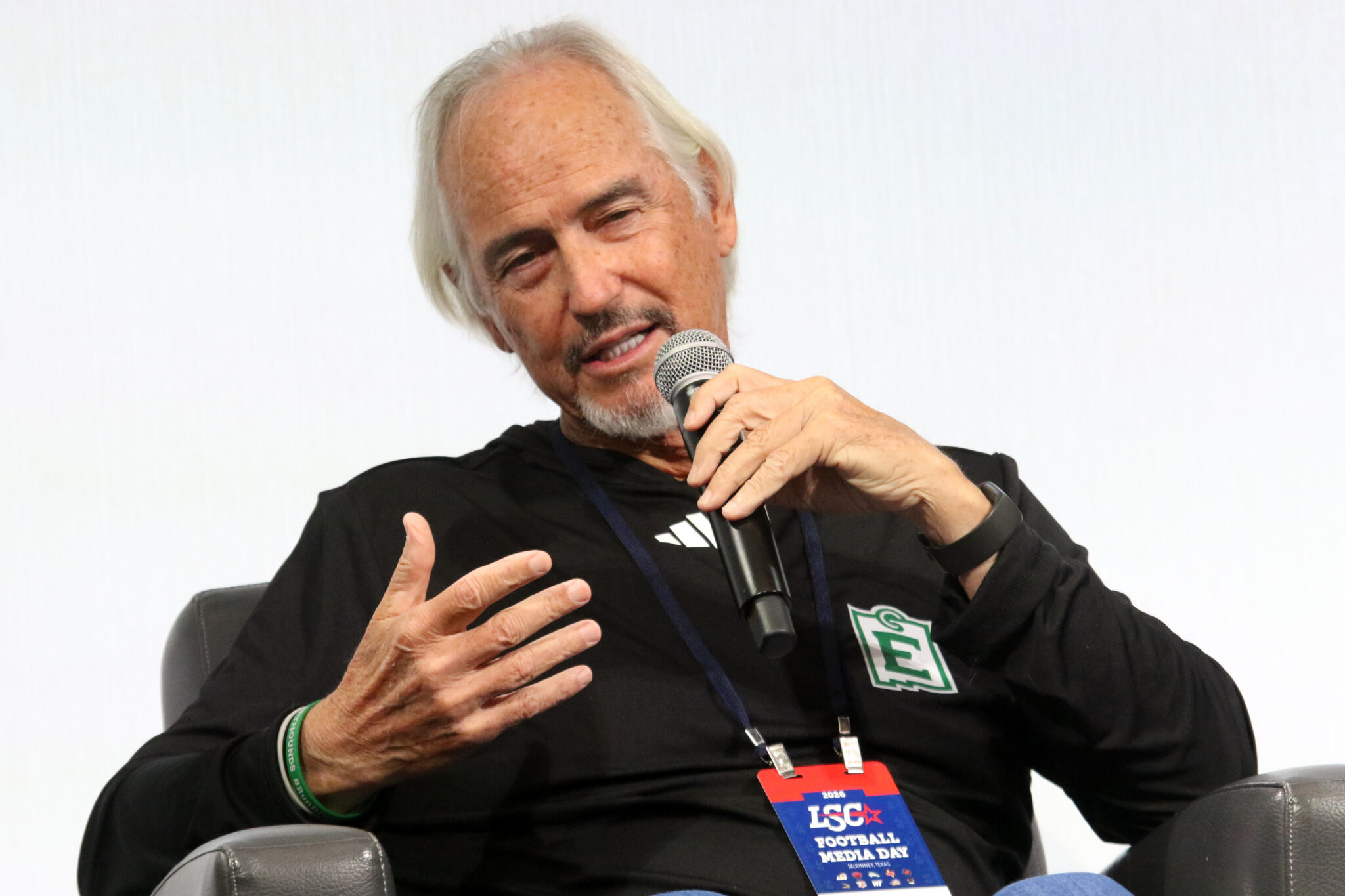 Art Briles - ENMU | Press Pass Sports Older man with white hair speaks into a handheld microphone during a panel discussion, wearing a black shirt and conference badge.