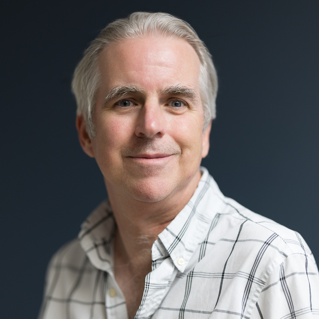 headshots-portraits-jpegJoe-3 | Press Pass Sports | Press Pass Sports Smiling older man with gray hair and a plaid white shirt against a dark blue background.