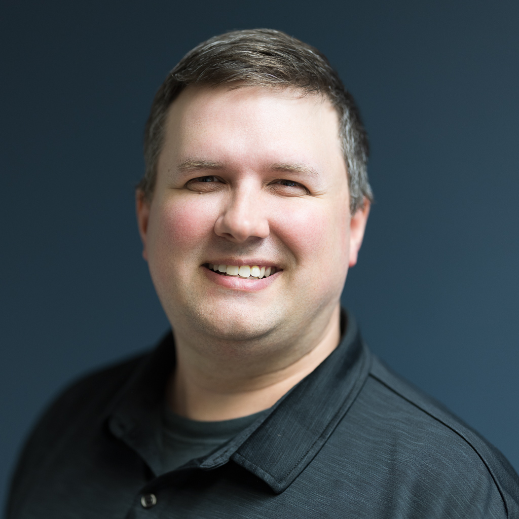 headshots-portraits-jpegJoe-2 | Press Pass Sports | Press Pass Sports Friendly head-and-shoulders portrait of a smiling man with short dark hair, wearing a black collared shirt, against a blue background.