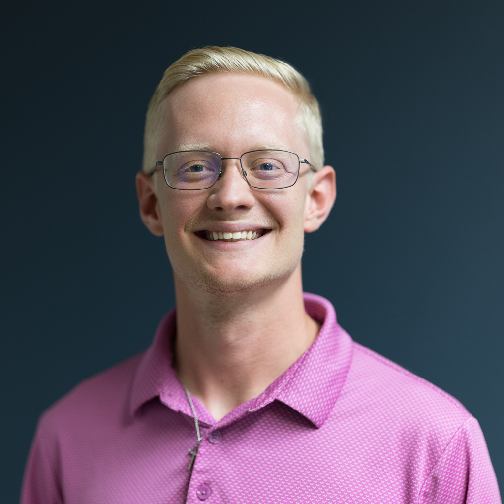 headshots-portraits-jpegJoe-1 | Press Pass Sports | Press Pass Sports Smiling man with short blond hair and glasses wearing a pink button-up polo against a dark blue background, posing for a portrait.