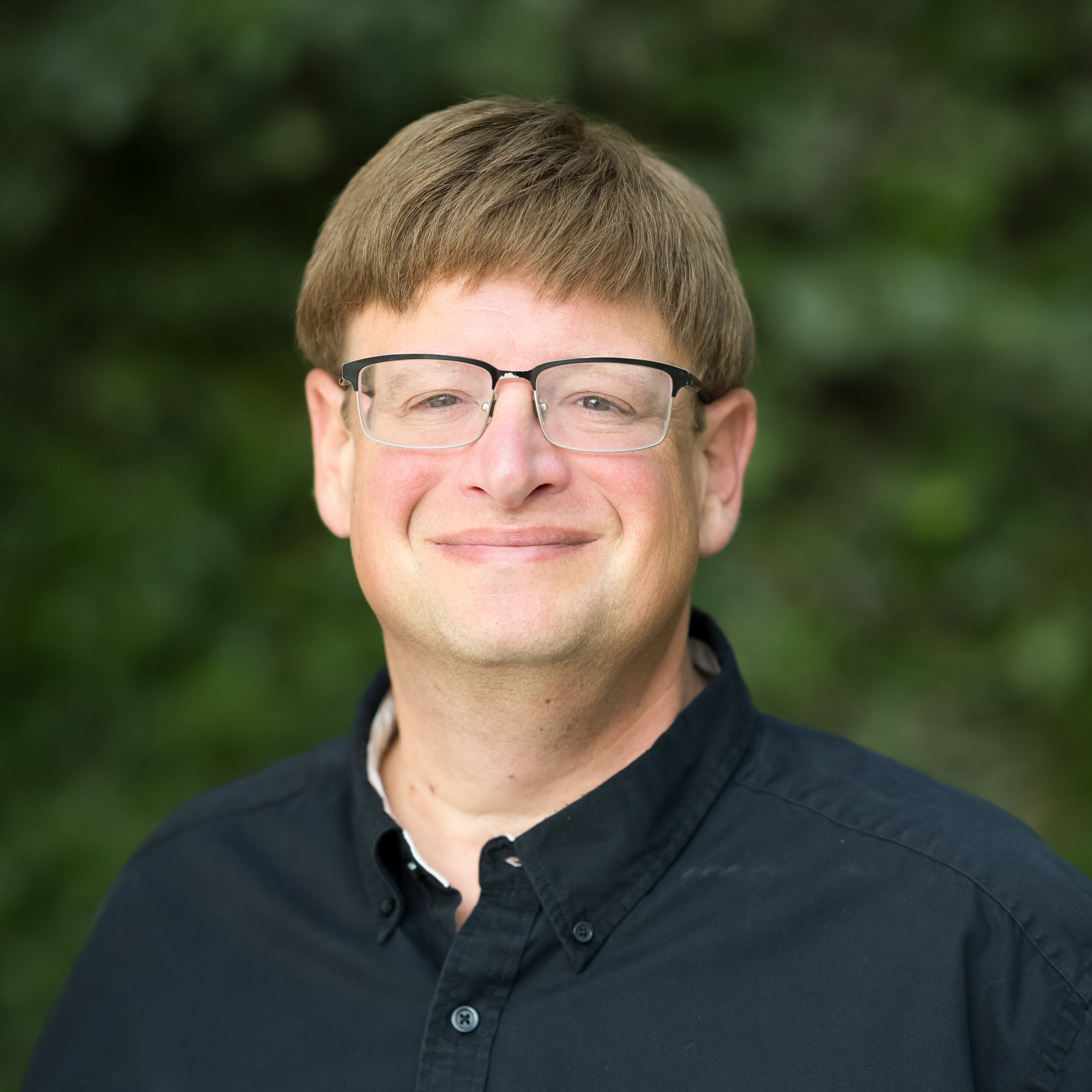 kale-headshot-jpegJoe-sq-1 | Press Pass Sports | Press Pass Sports Close-up portrait of a man wearing rectangular glasses and a dark button-down shirt, outdoors with a green blurred background.