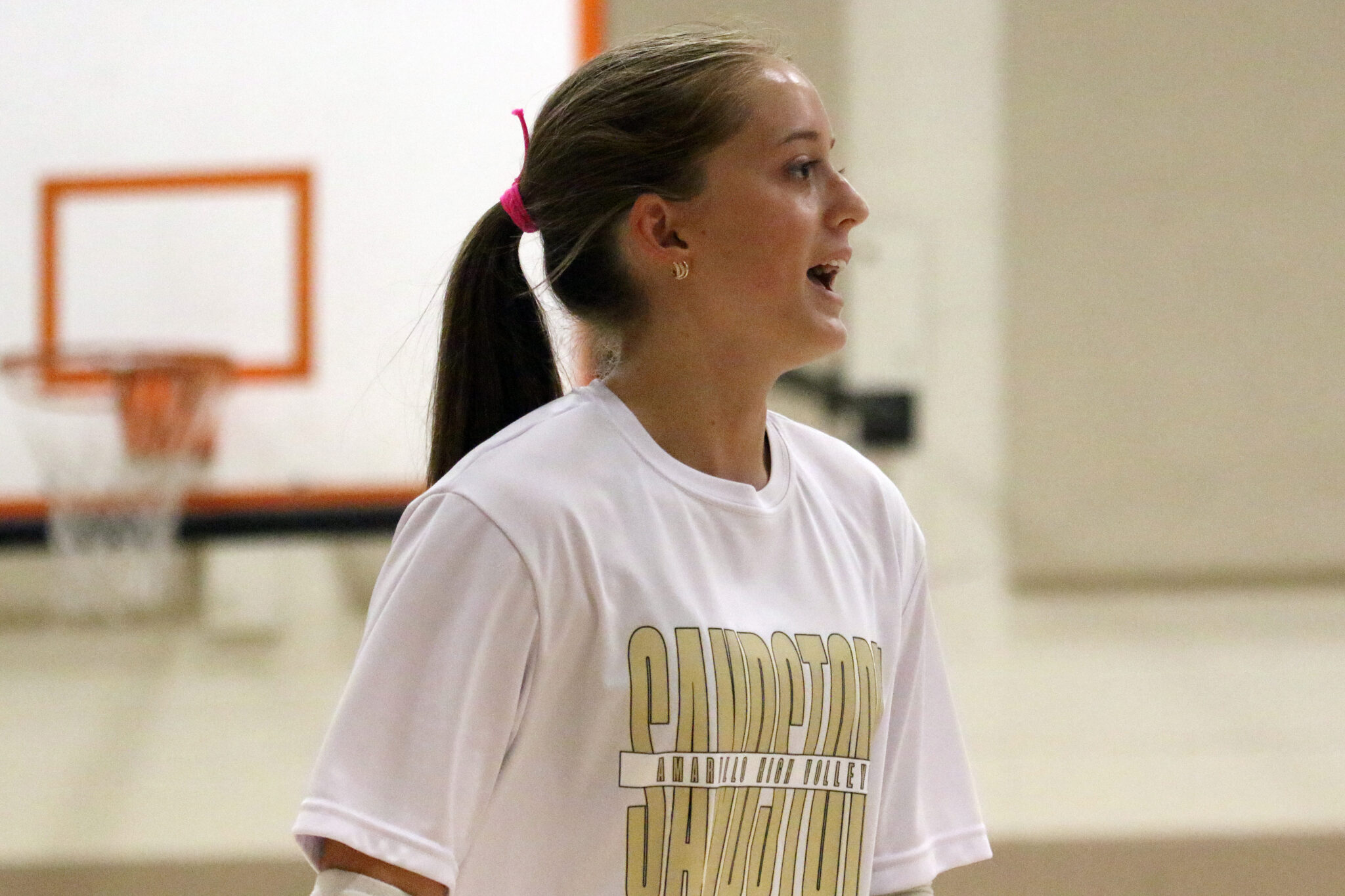 1K6A4799 | Press Pass Sports Side profile of a young woman with a ponytail wearing a white T‑shirt in a gym, a basketball hoop blurred in the background.