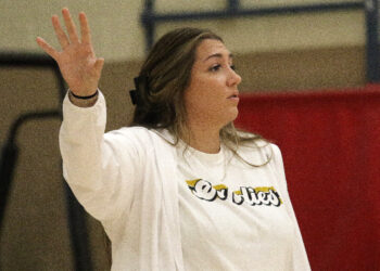 1K6A4889 | Press Pass Sports Woman in a white jacket raises her left hand in the air, facing to the right in a gym-like setting.