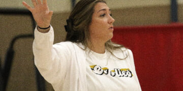 Woman in a white jacket raises her left hand in the air, facing to the right in a gym-like setting.