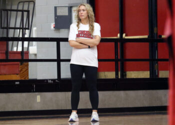 1K6A5159 | Press Pass Sports Woman with long blonde hair stands with arms crossed in a gym, wearing a white volleyball shirt and black leggings.