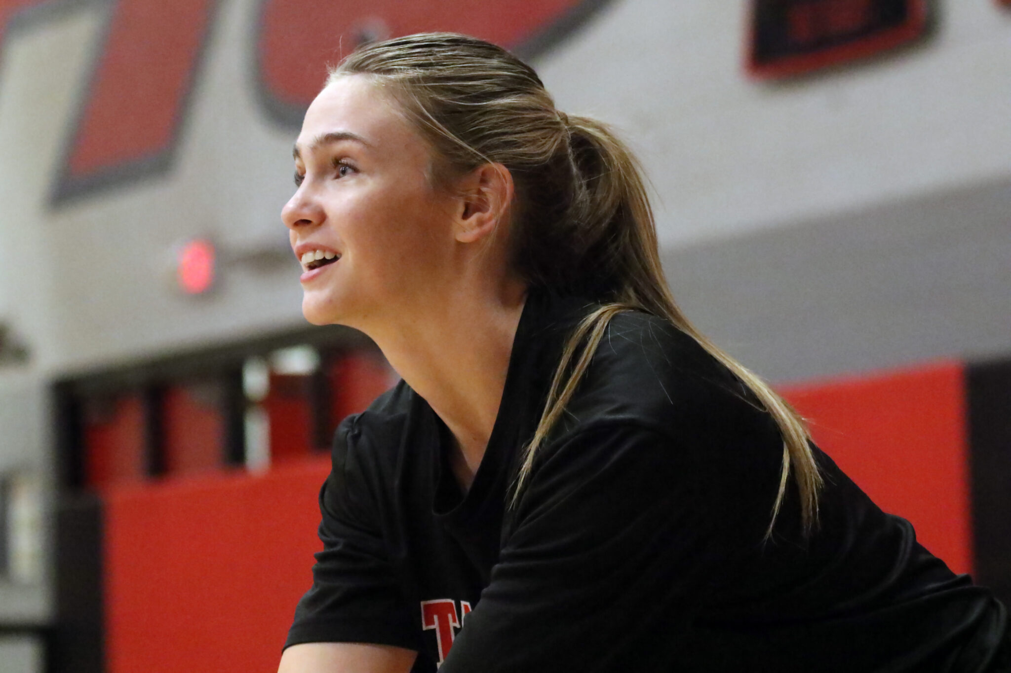 Woman in a gym, side profile with blonde ponytail, wearing a black top, captured in mid-action against a red and gray background.