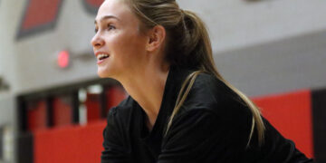 Woman in a gym, side profile with blonde ponytail, wearing a black top, captured in mid-action against a red and gray background.