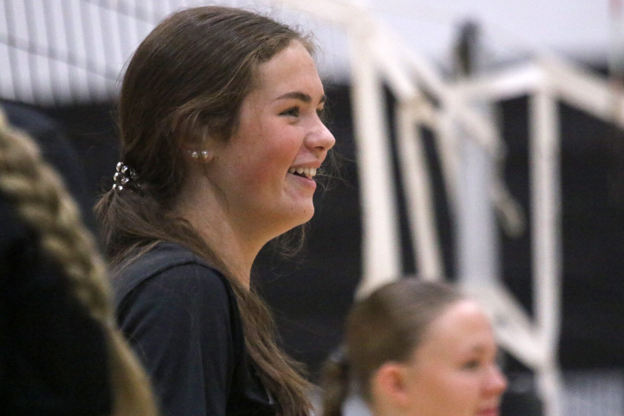 1K6A4234 | Press Pass Sports Side profile of a smiling young woman with long brown hair, indoors among a crowd.