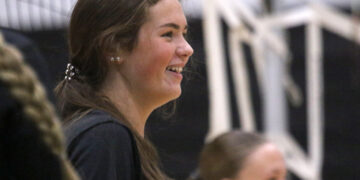 Side profile of a smiling young woman with long brown hair, indoors among a crowd.