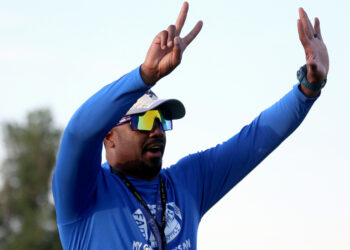Palo Duro - Eric Mims 1 | Press Pass Sports Athlete in a blue long-sleeve shirt raises a two-finger victory sign with one hand, wearing a cap, sunglasses, and a lanyard outdoors.
