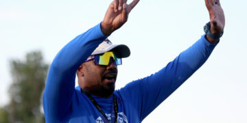 Athlete in a blue long-sleeve shirt raises a two-finger victory sign with one hand, wearing a cap, sunglasses, and a lanyard outdoors.